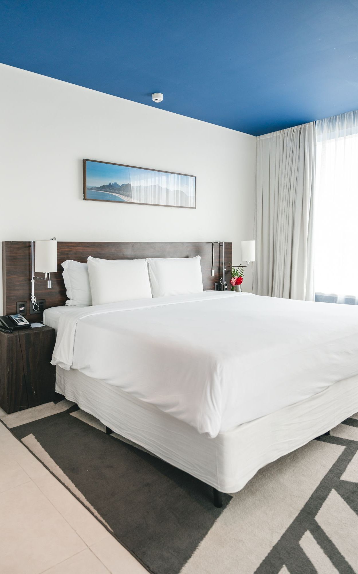 Um quarto de hotel bem iluminado com uma cama grande e branca, cabeceira de madeira escura e teto azul. Um quadro está pendurado acima da cama.