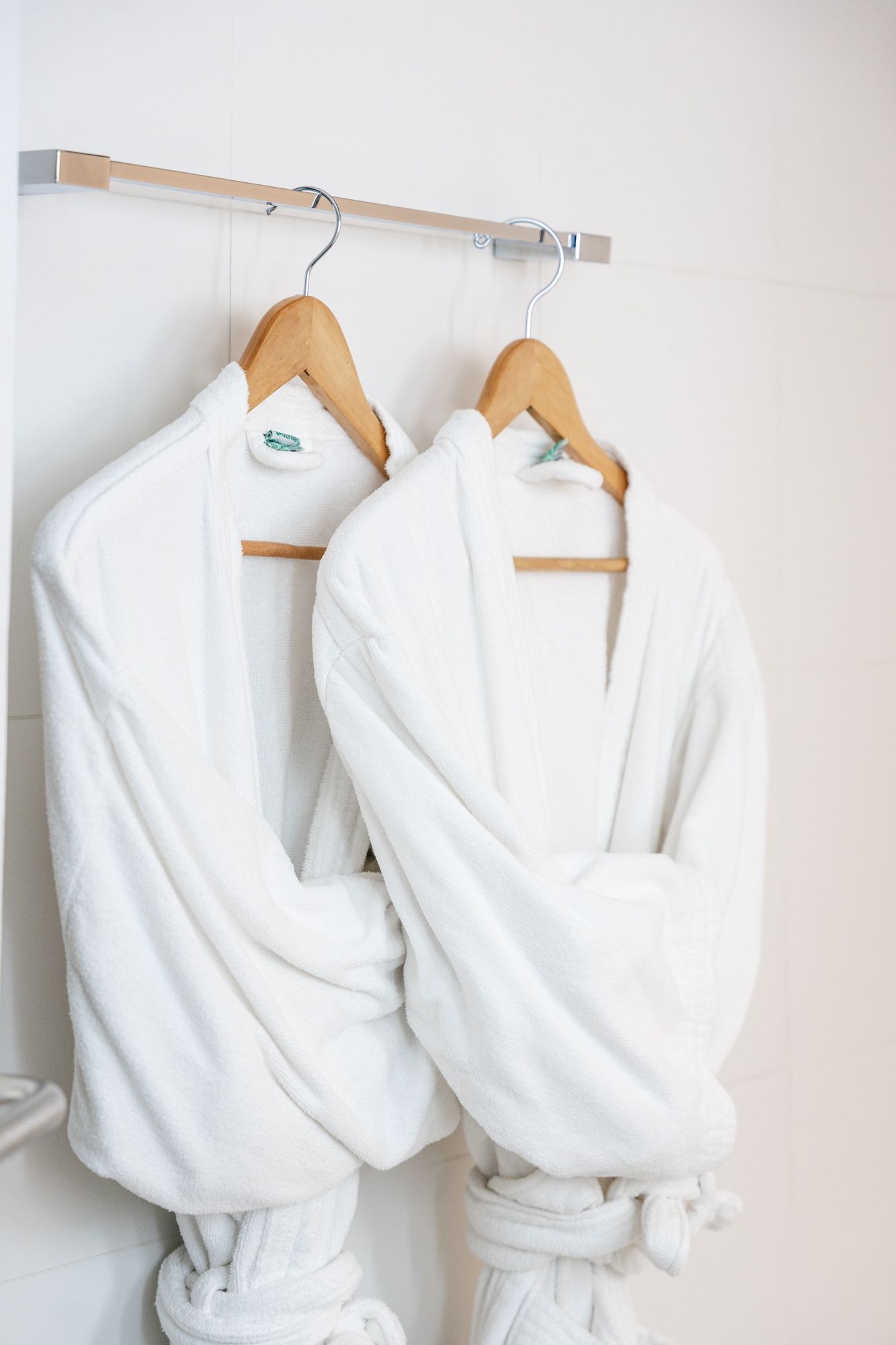 Dois roupões brancos pendurados em cabides de madeira em um cabide prateado contra uma parede branca.