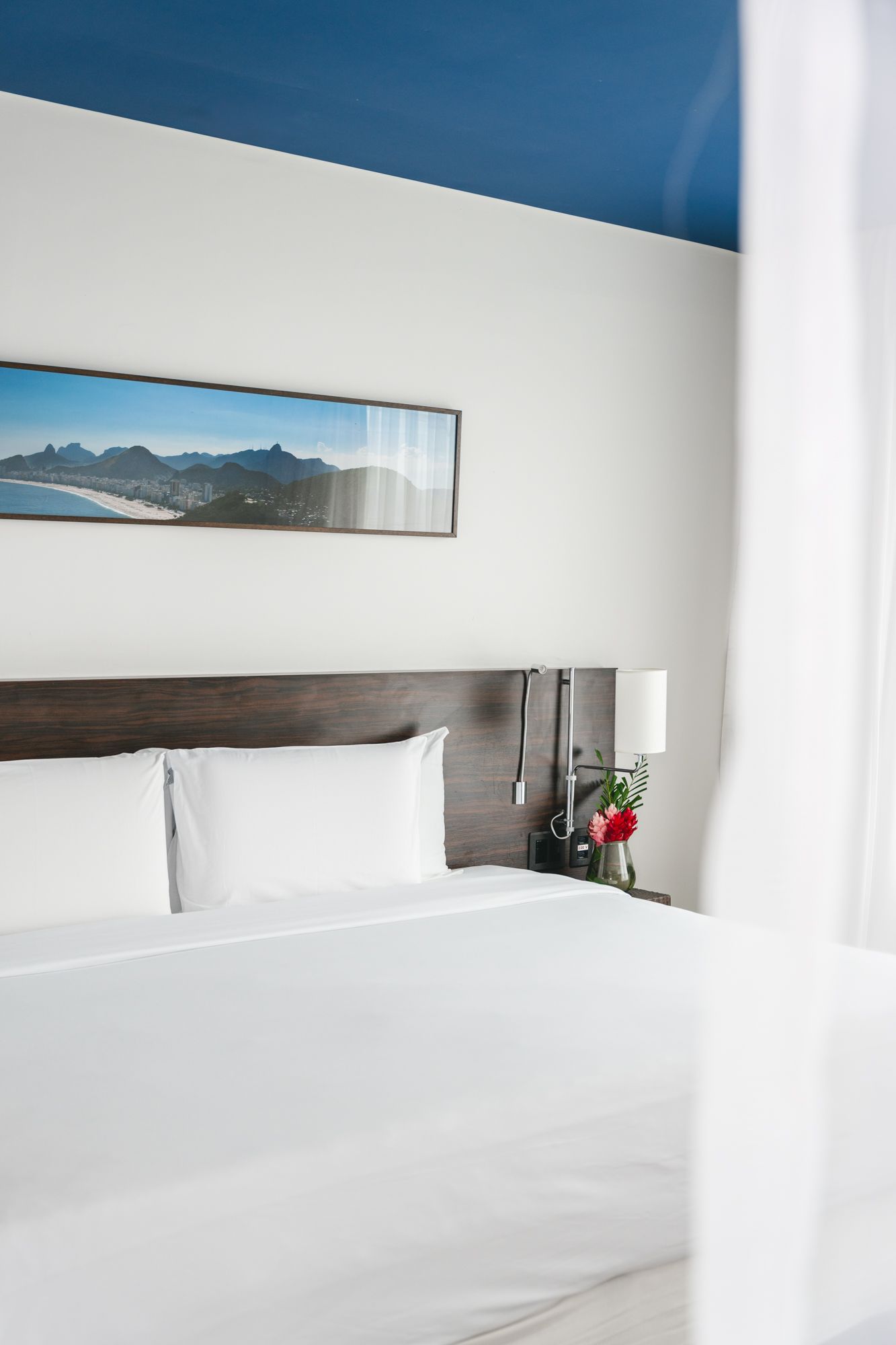 Um quarto de hotel moderno com uma cama king-size, lençóis brancos e teto azul; uma pintura de paisagem pendurada acima da cabeceira.