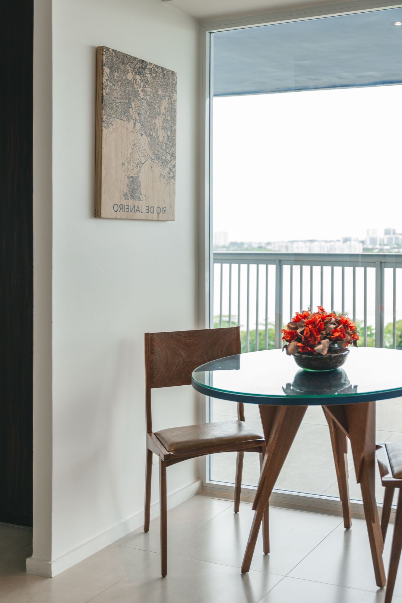 Uma pequena sala de jantar com uma mesa redonda de vidro, cadeiras de madeira e uma janela com vista para uma varanda. Um mapa pendurado na parede.