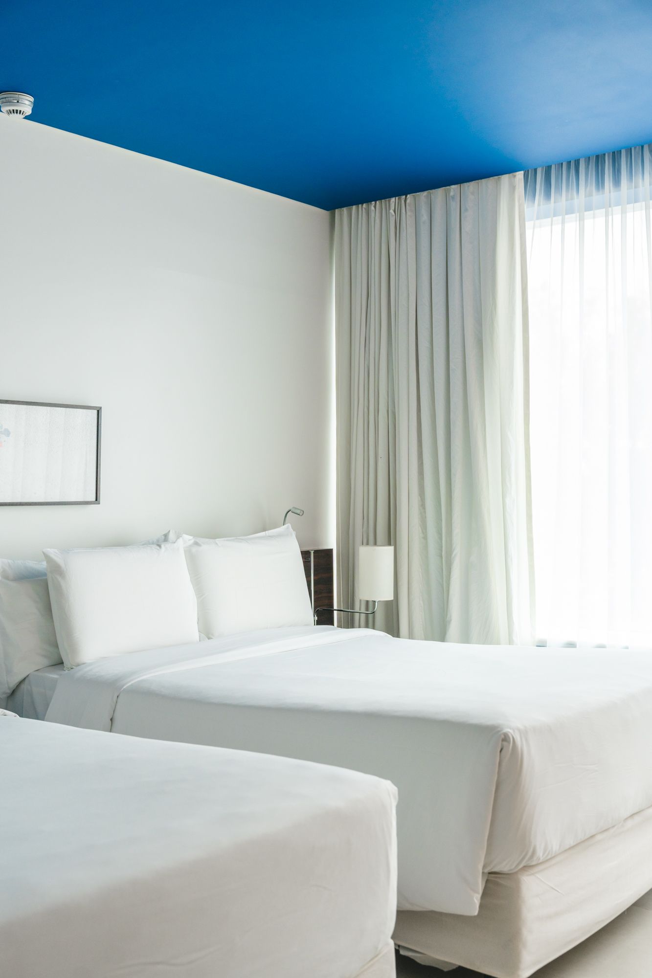 Quarto de hotel com paredes brancas, duas camas brancas, cortinas transparentes e teto azul.