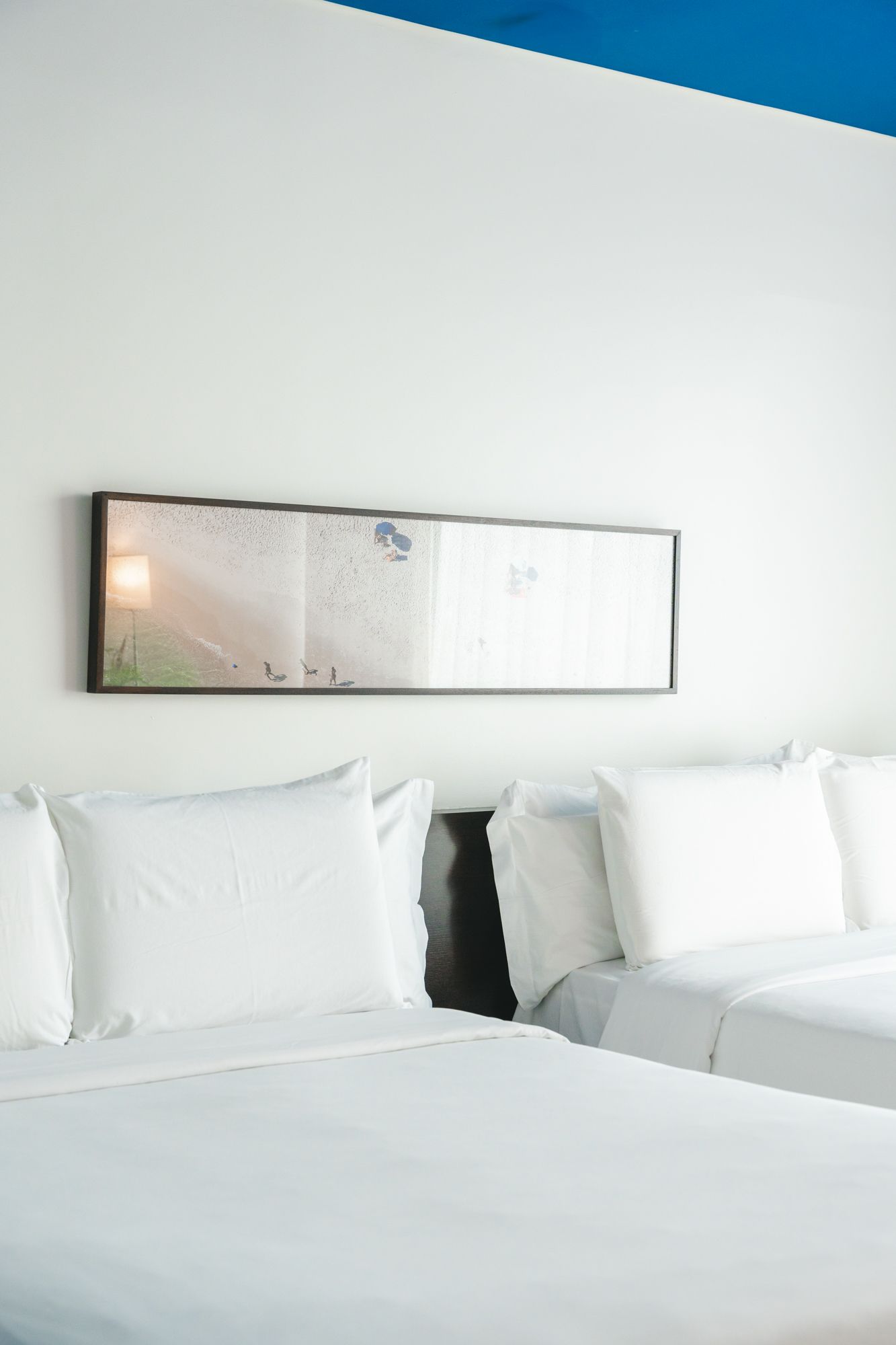 Duas camas com roupa de cama branca estão em um quarto. Uma obra de arte emoldurada está pendurada acima das camas. A parede é branca com um tampo azul.