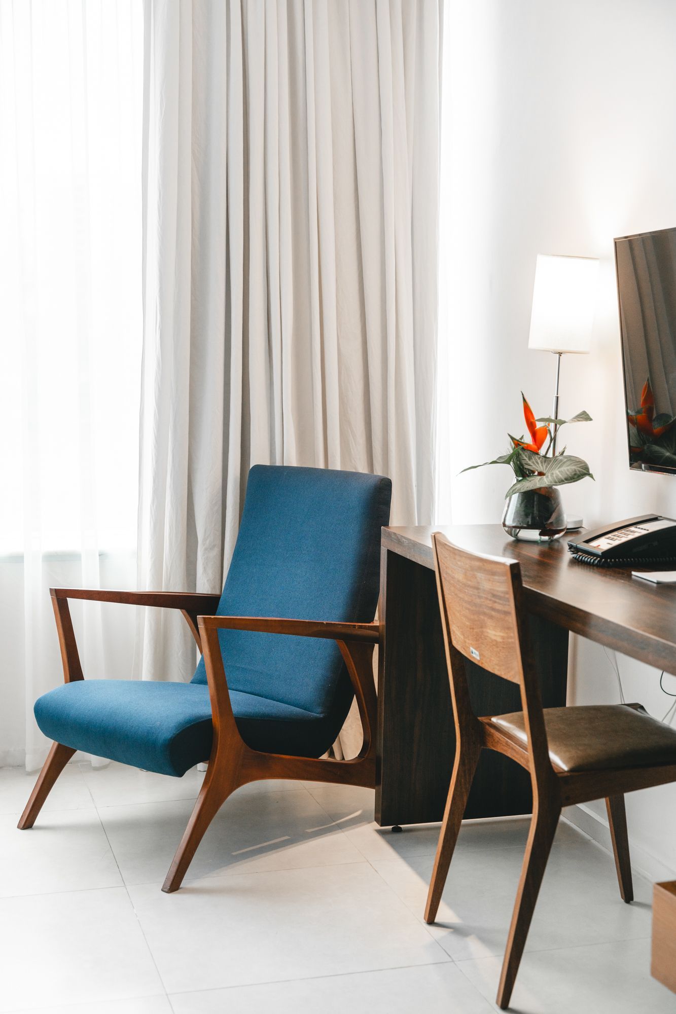 Uma poltrona azul ao lado de uma mesa e cadeira de madeira em uma sala com uma janela e cortinas brancas.