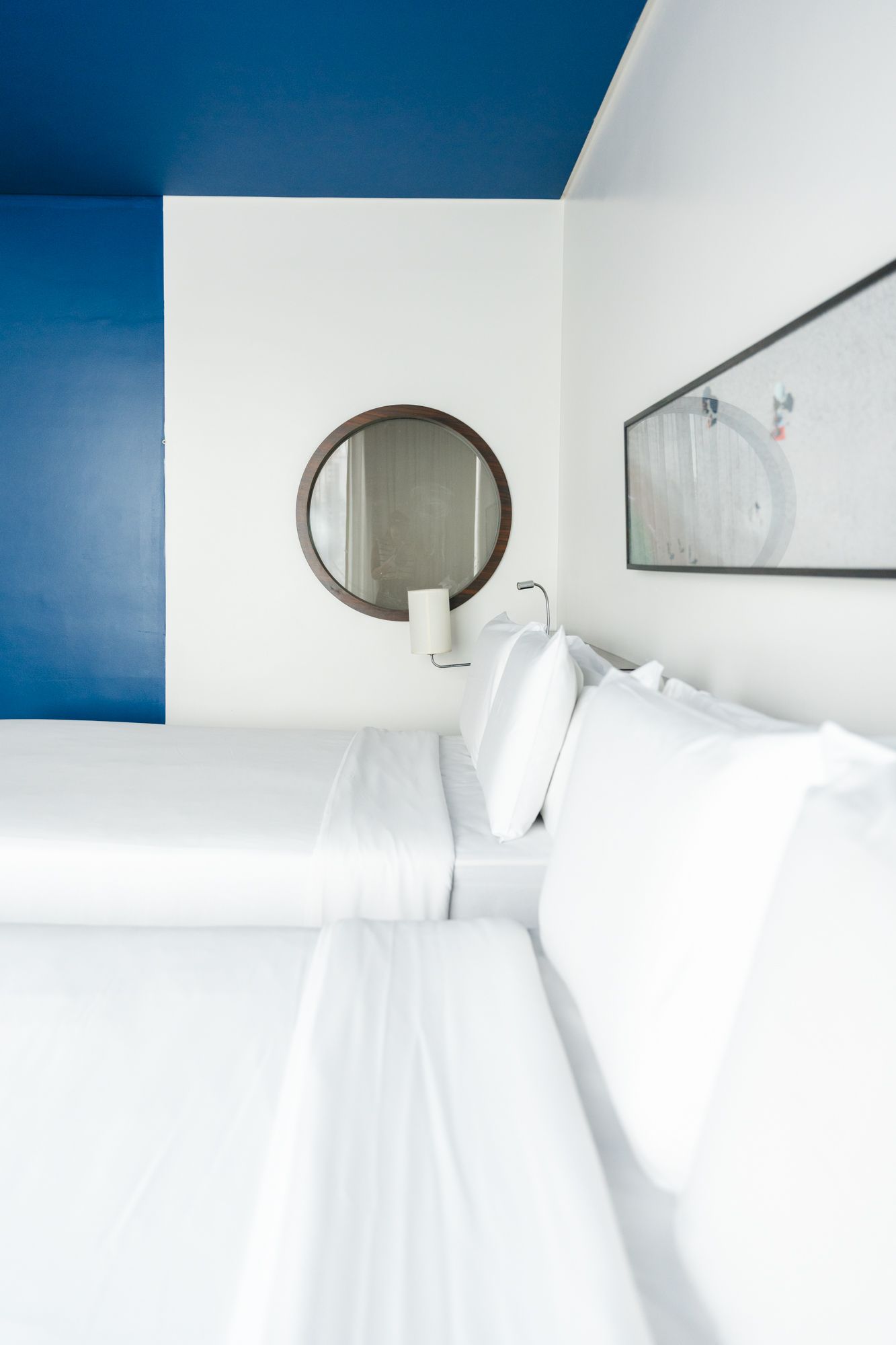 Um quarto de hotel moderno com duas camas brancas, um espelho redondo e um esquema de cores azul e branco.