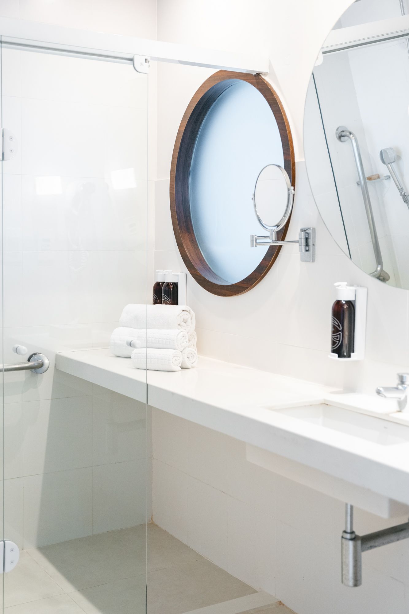 Banheiro branco com espelho oval, bancada e chuveiro atrás de vidro. Toalhas brancas empilhadas sobre o balcão.