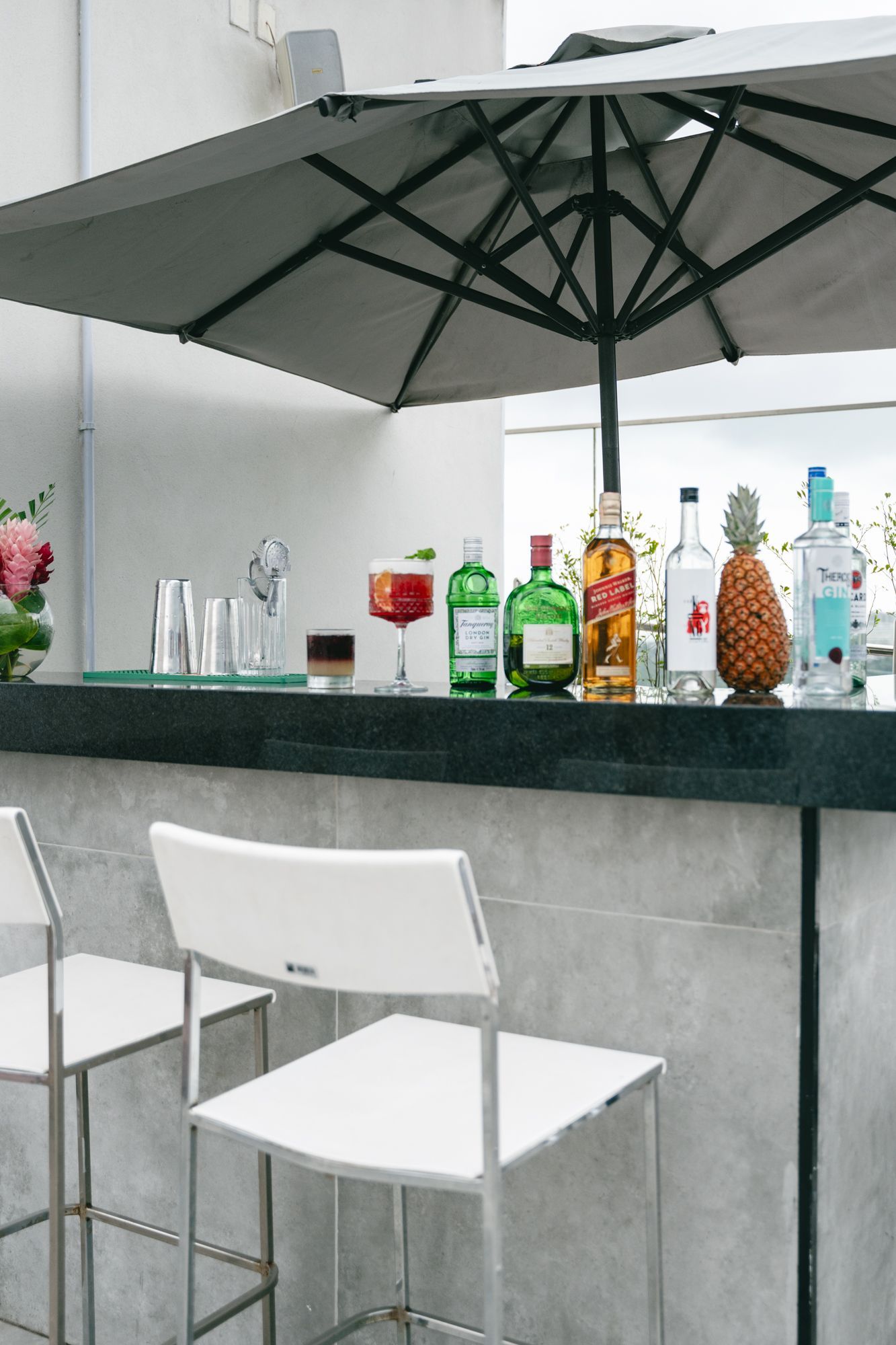 Bar ao ar livre com cadeiras brancas, coquetéis e garrafas sob um guarda-sol cinza.