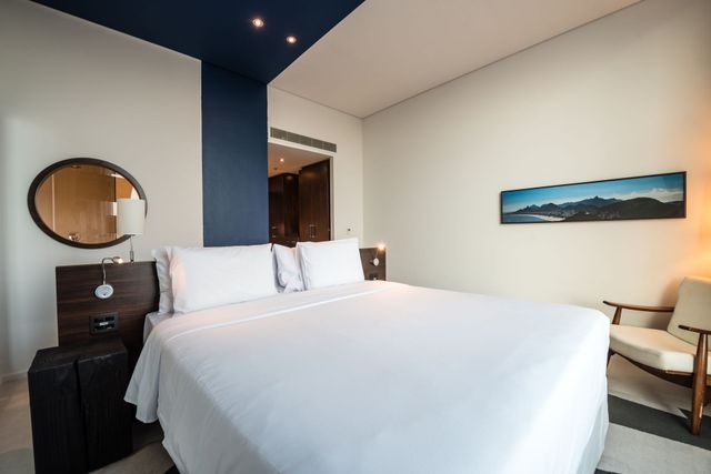 Um quarto de hotel moderno com cama, cadeira e obras de arte. Detalhes em azul contrastam com paredes brancas e móveis de madeira.