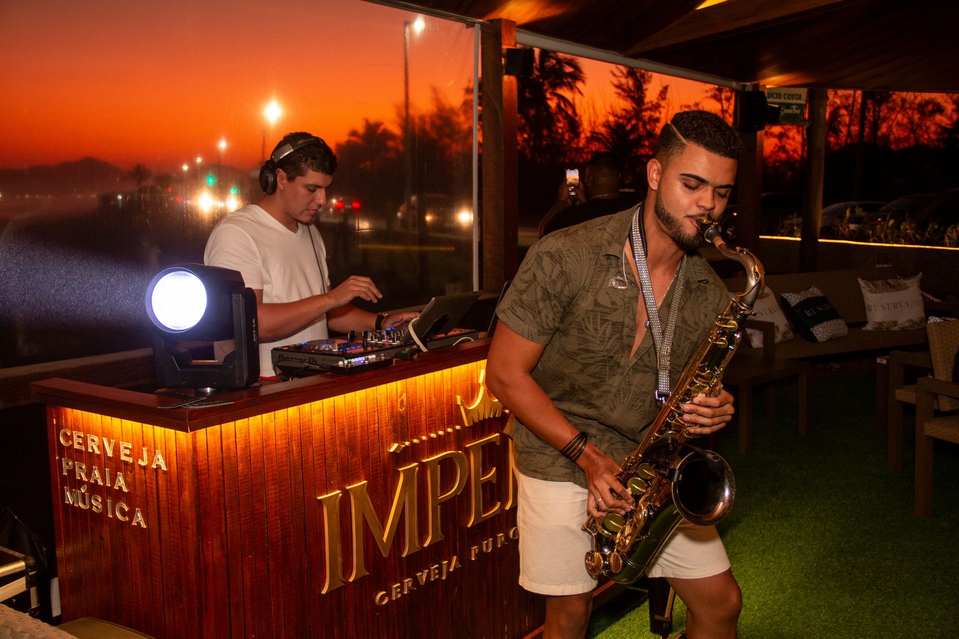 DJ e saxofonista se apresentando em um bar de praia ao pôr do sol. O bar tem uma fachada de madeira com o nome 