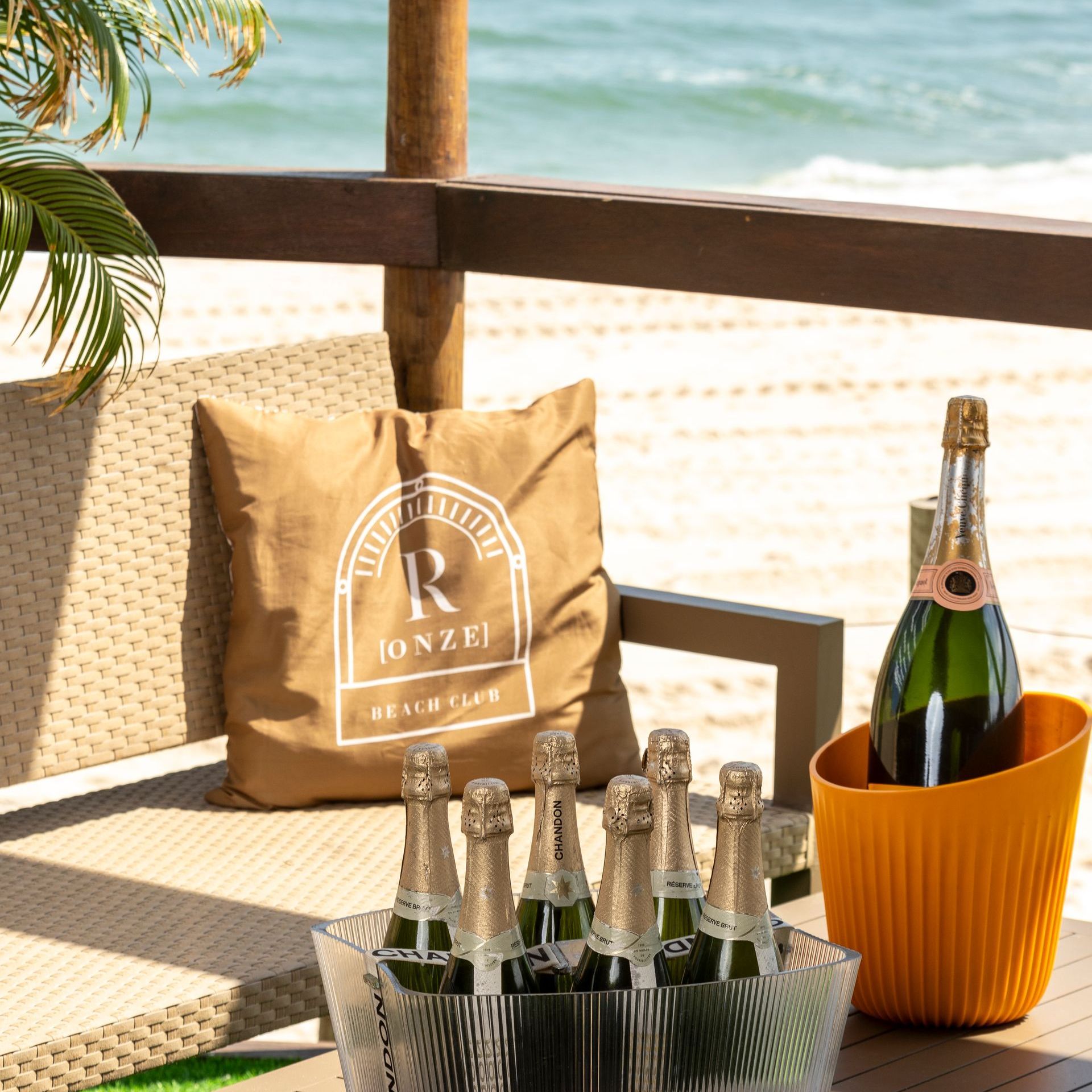 Garrafas de champanhe gelando sobre uma mesa com vista para a praia. Uma garrafa está dentro de um balde de gelo laranja. Uma almofada em uma cadeira exibe o logotipo 