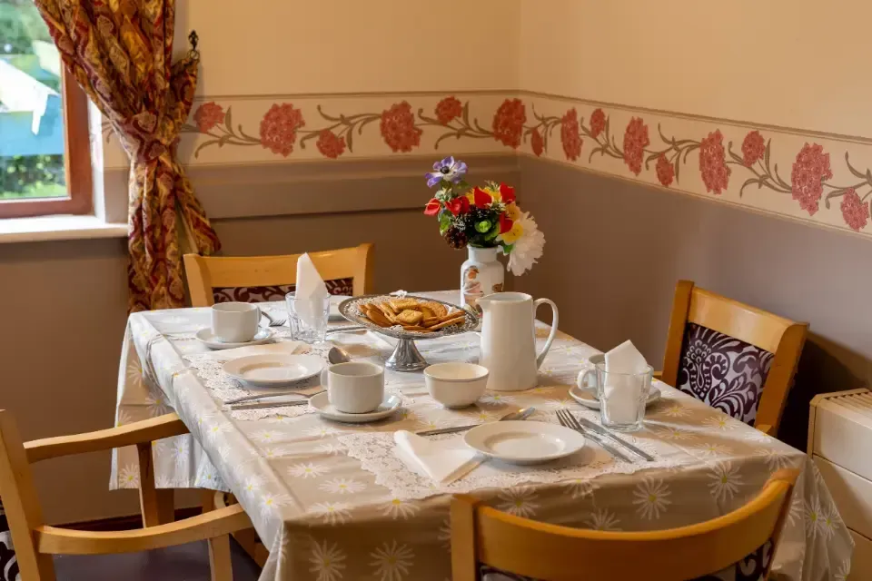 Oaklands Residential Care Home, Methley, Leeds | A table with plates , cups , and a vase of flowers on it in a dining room.