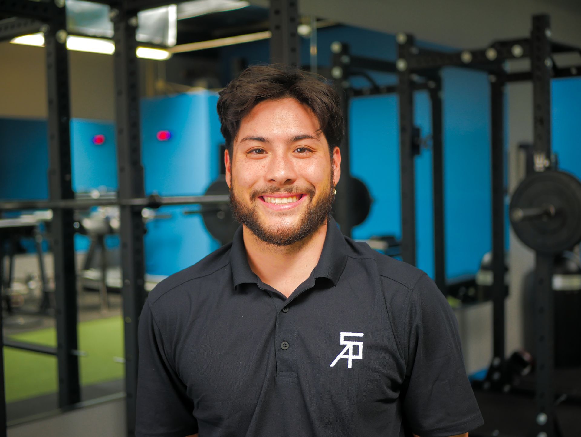 Smiling man in black shirt, in a gym, with workout equipment behind him.