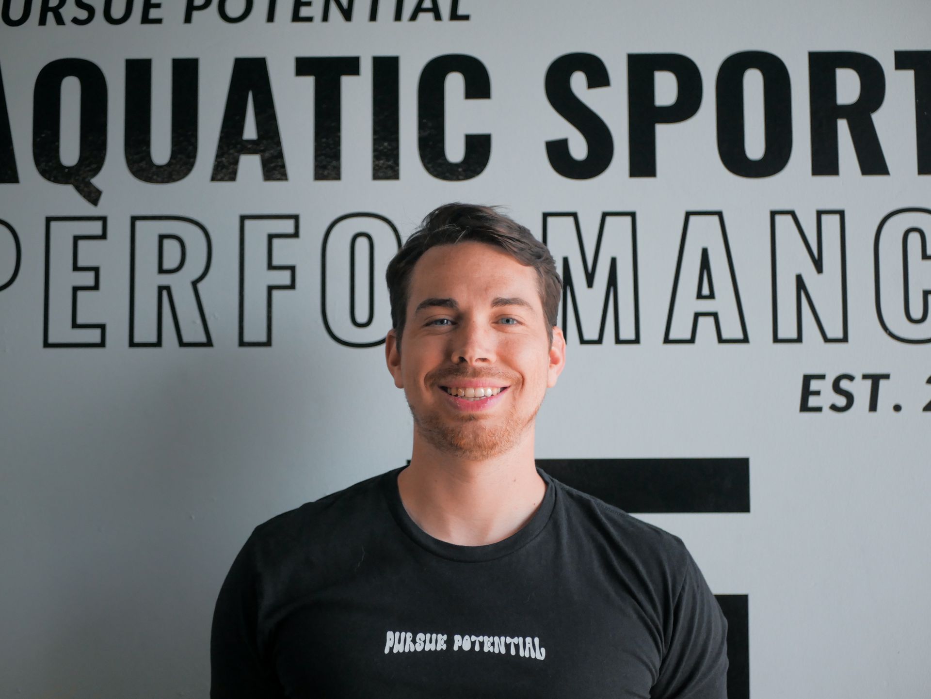 Man smiling in front of Aquatic Sport Performance sign. He wears a black t-shirt.