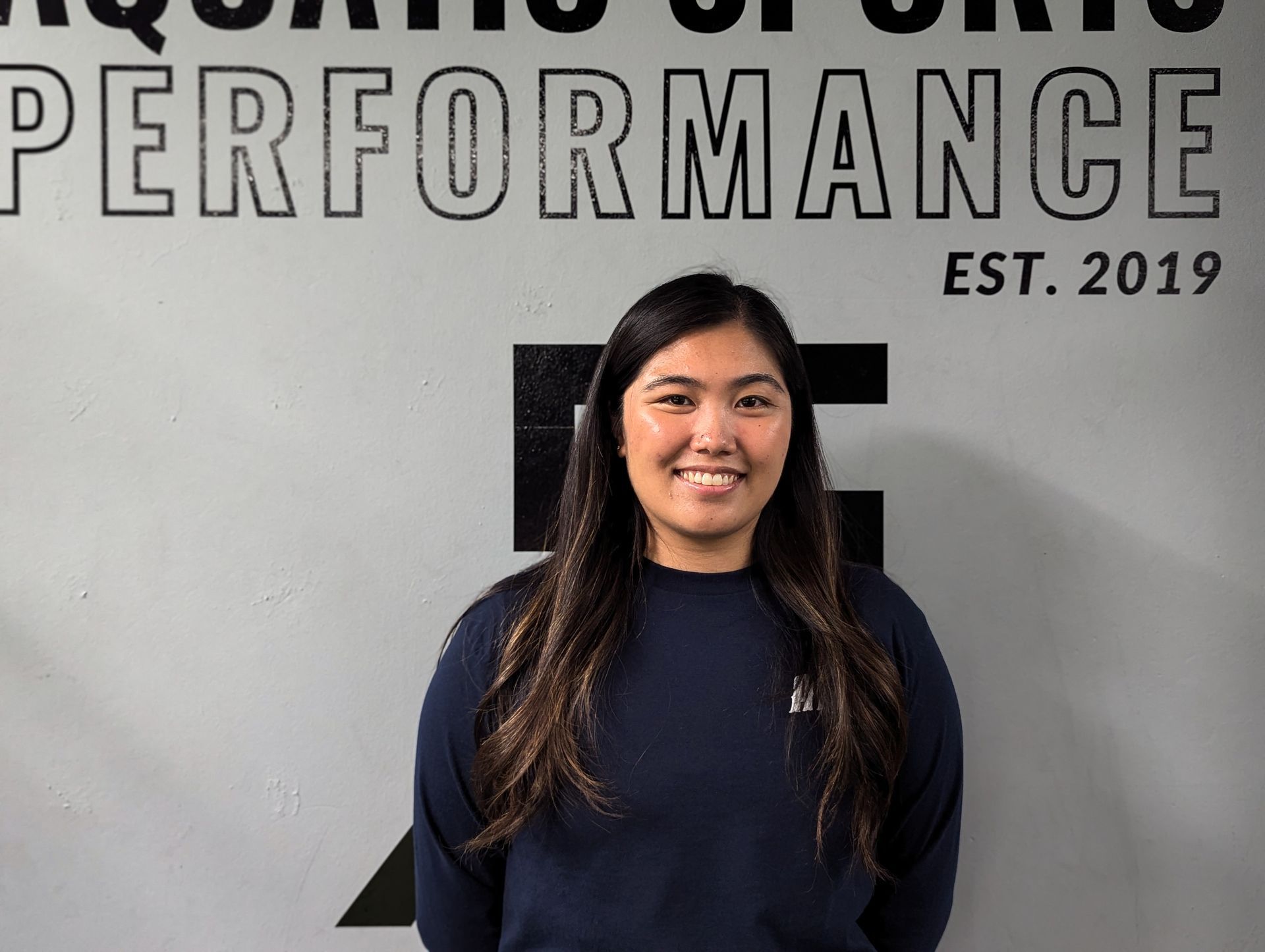 Young woman smiling, wearing a navy blue sweater, standing in front of a grey wall with black text.