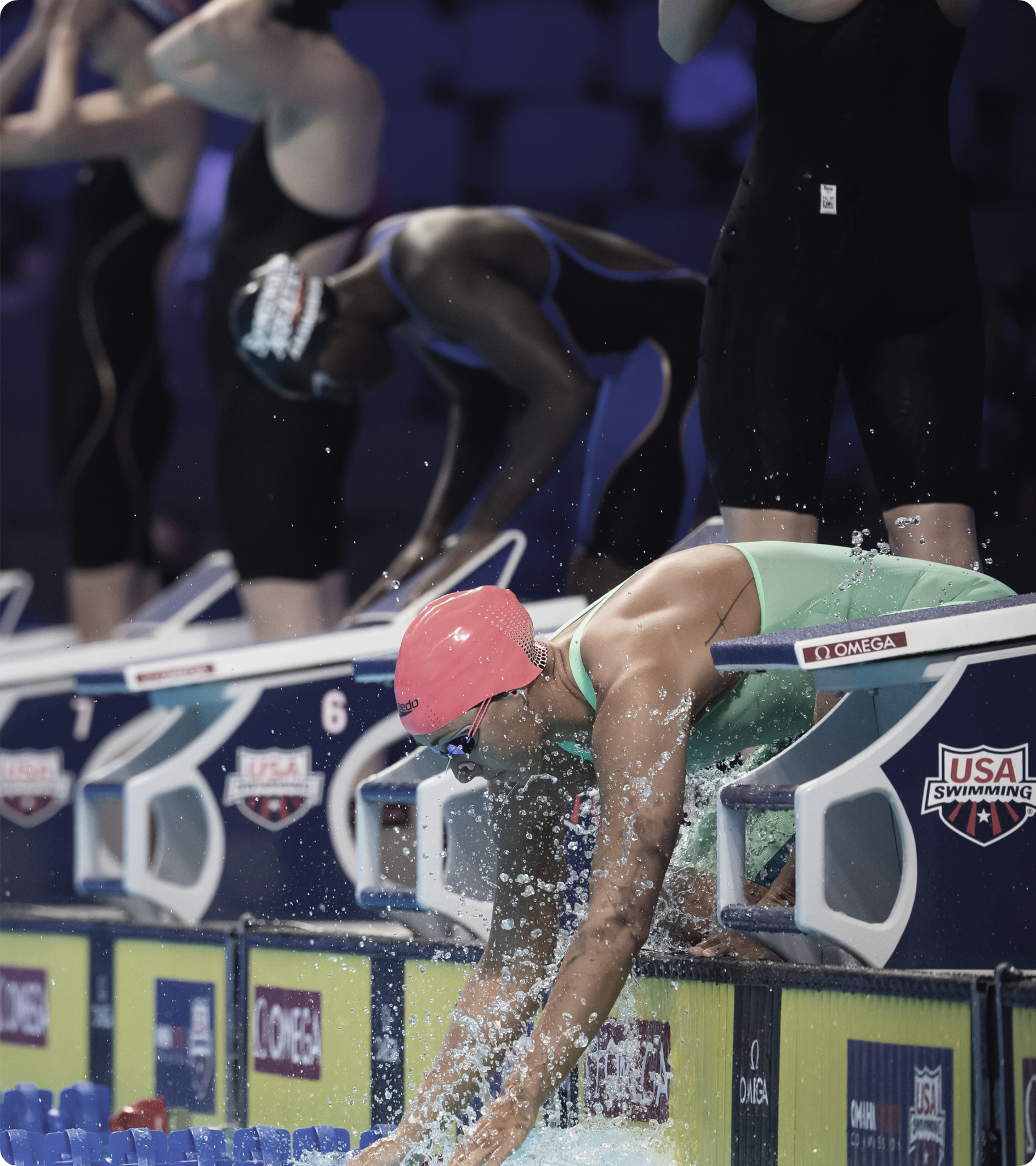 Swimmers in their starting positions at a competition. One swimmer with pink cap dives in the pool.