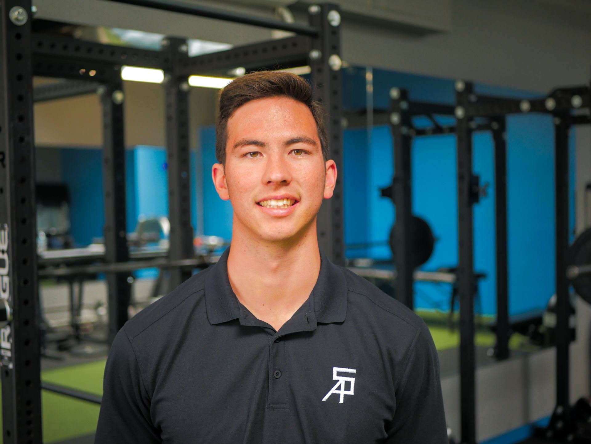 Young man in a black polo shirt smiles in a gym setting.