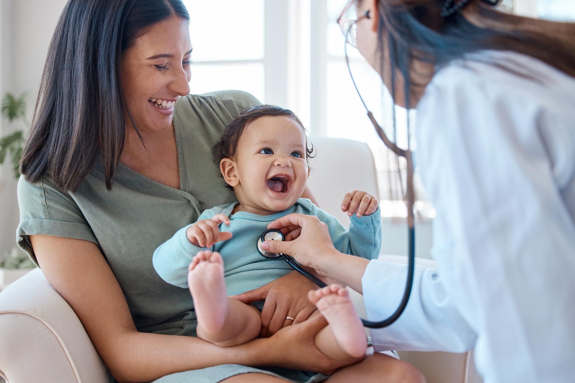 Smiling mother holding a laughing baby while a pediatrician uses a stethoscope during a clinic visit