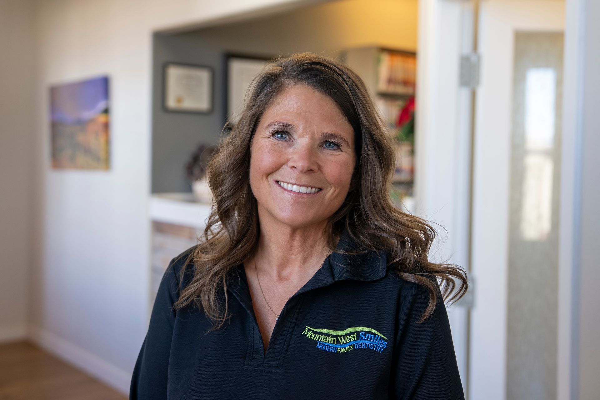 Woman with brown hair, smiling, wearing black scrubs, a small necklace, and name tag.