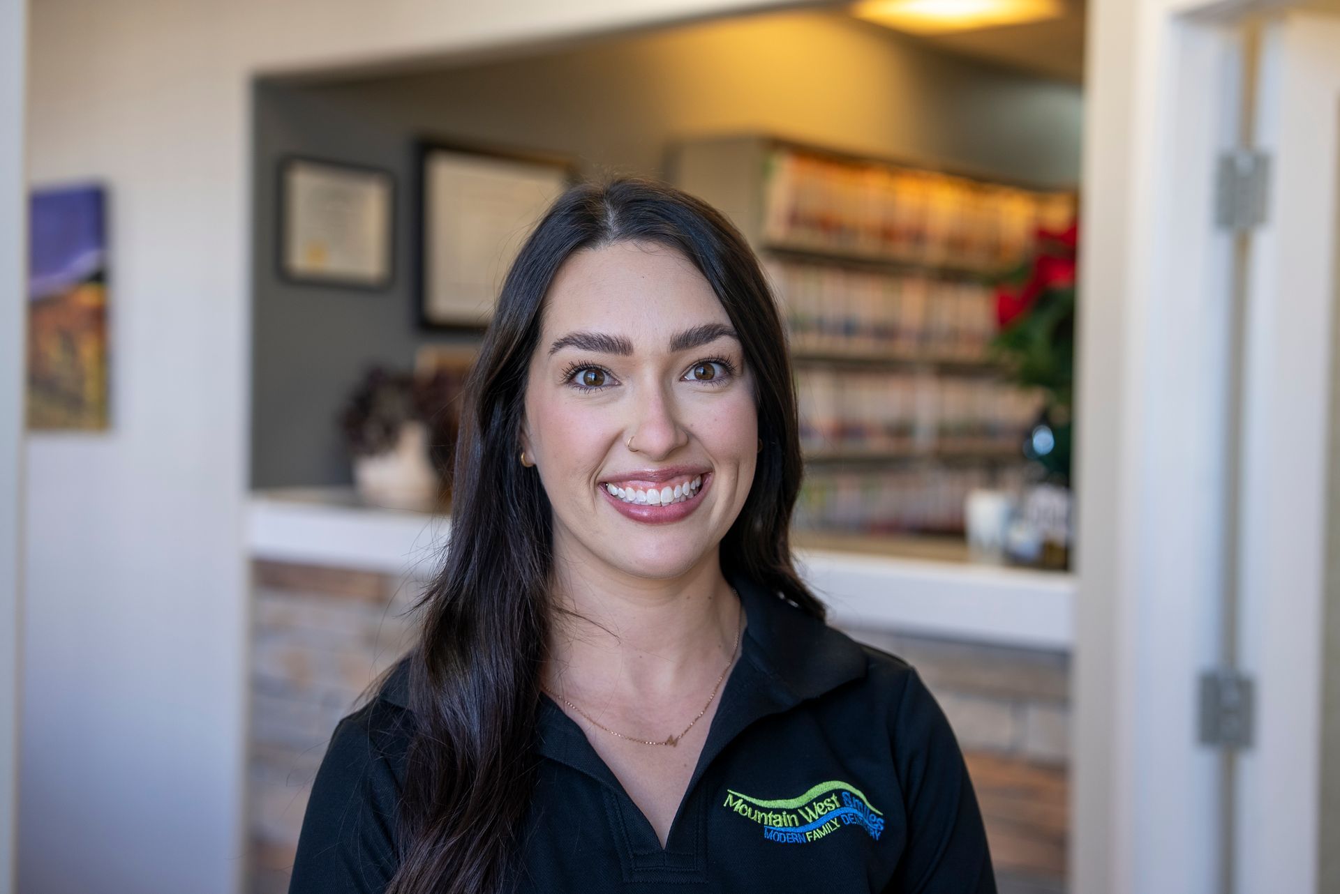 Woman smiling, wearing black scrubs, red-framed glasses, in a dental office setting.