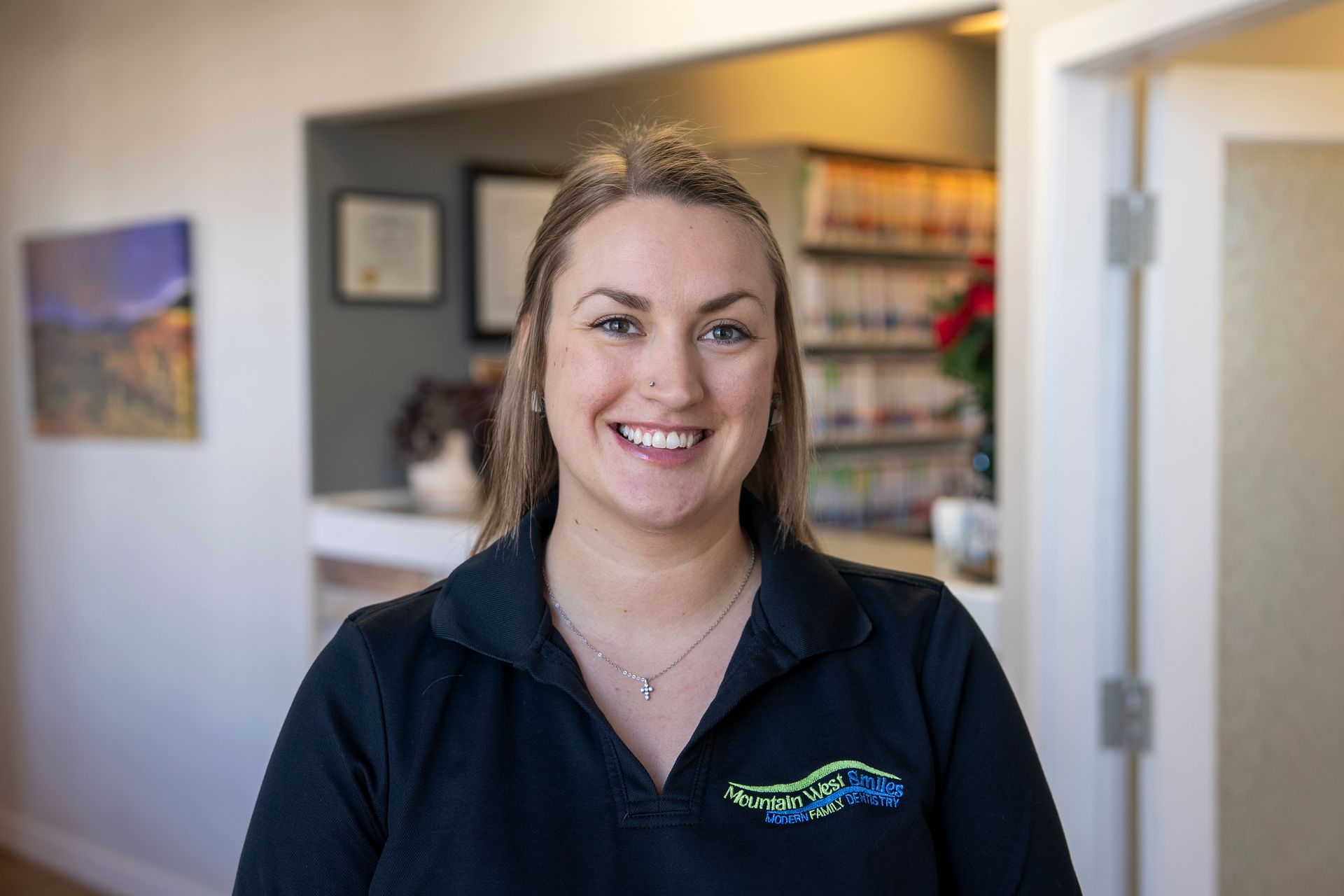 Woman with long blonde hair smiles, wearing a burgundy sweater indoors. A clock is visible on the wall behind her.