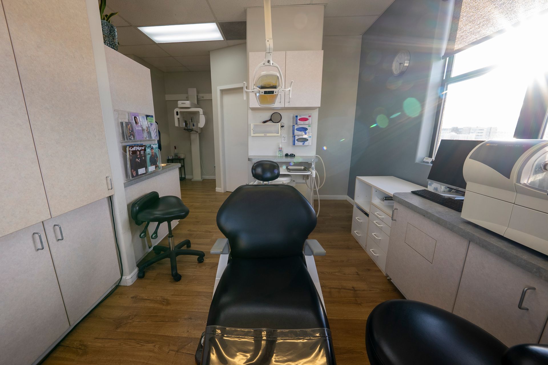 Dental operatory with patient chair, cabinets, computer, and x-ray display on wall.