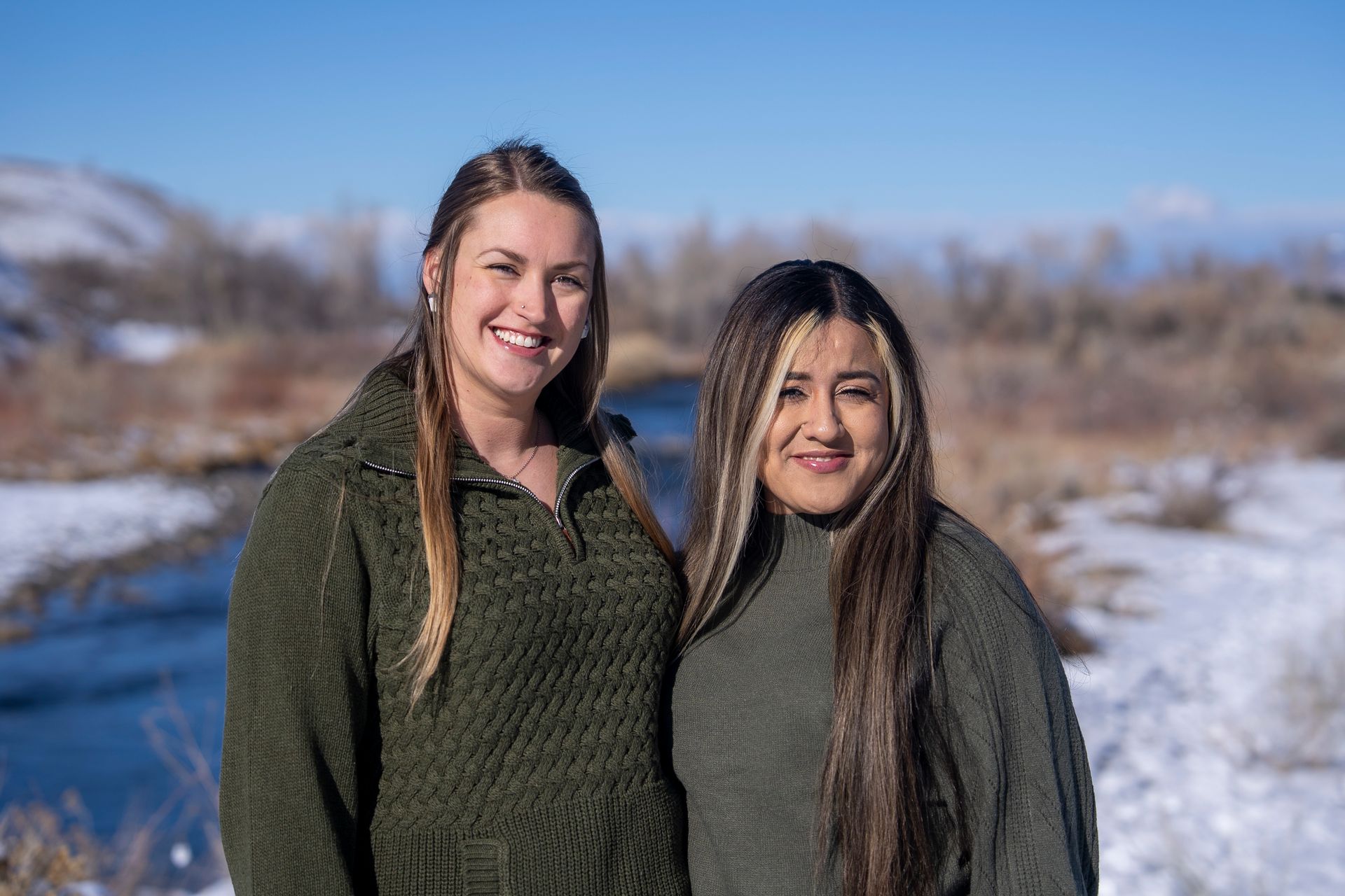 Two women smiling, standing next to each other outdoors near a river with snow on the ground.