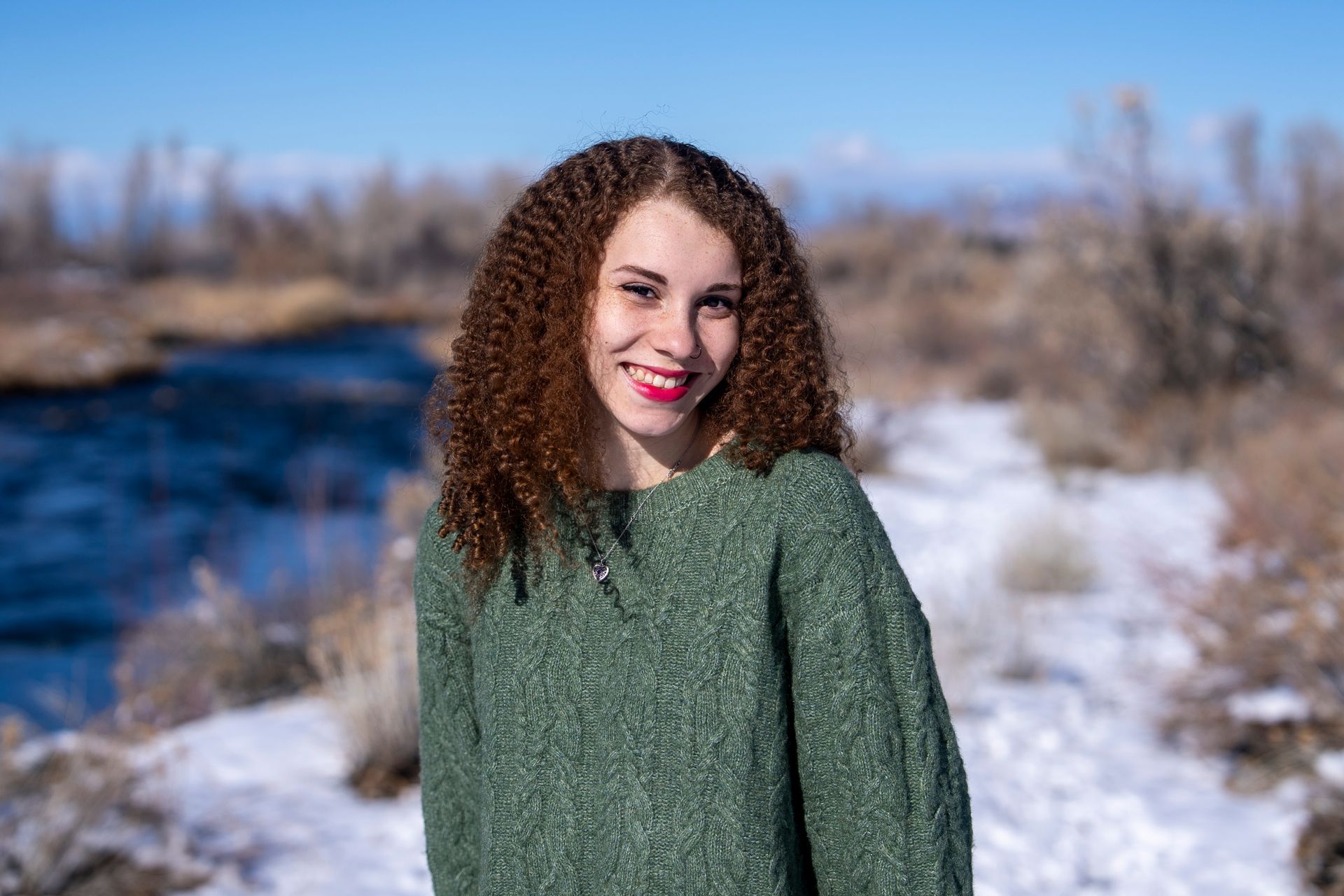 Woman smiling, wearing a green sweater, standing near snow and a river.