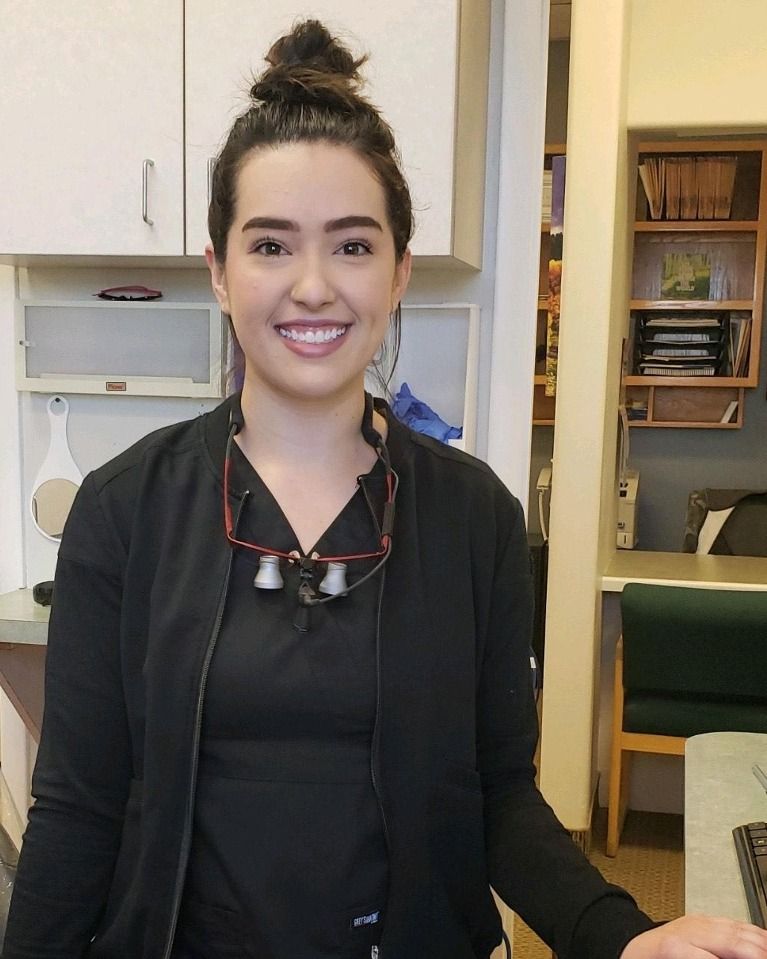 Woman smiling, wearing black scrubs, red-framed glasses, in a dental office setting.