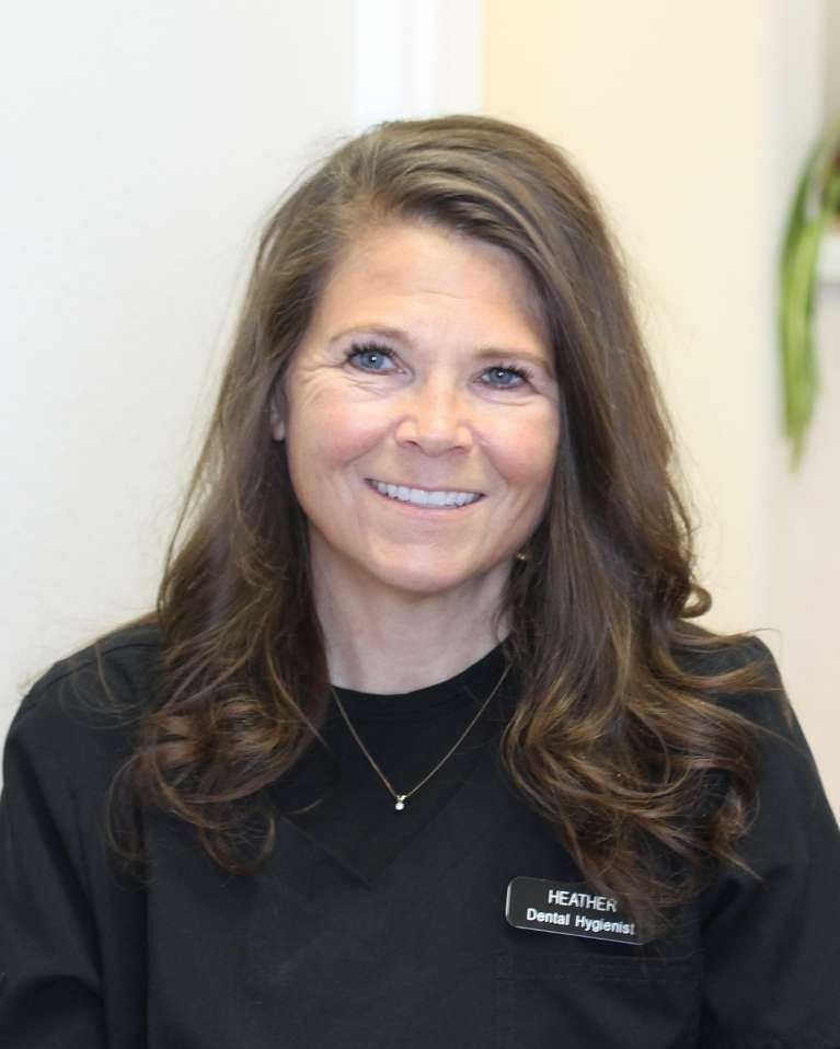 Woman with brown hair, smiling, wearing black scrubs, a small necklace, and name tag.