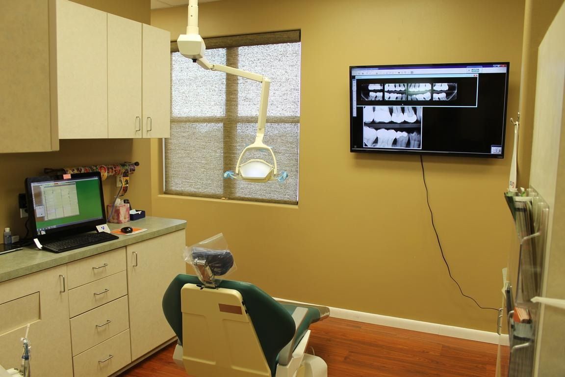 Dental operatory with patient chair, cabinets, computer, and x-ray display on wall.