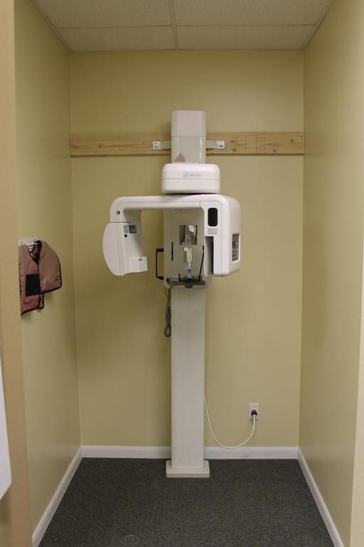 A white panoramic X-ray machine in a small dental office room, beige walls, gray floor.