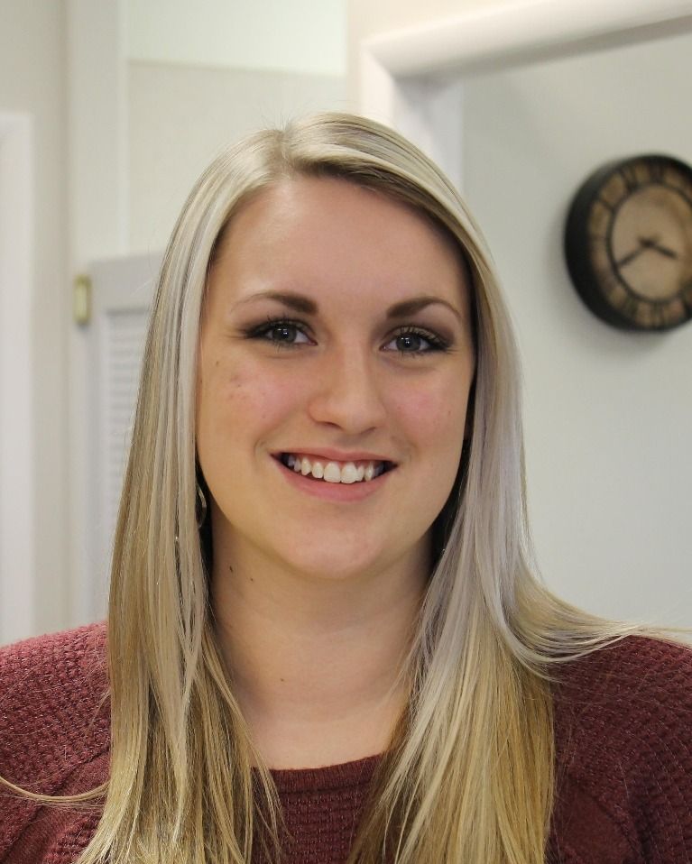 Woman with long blonde hair smiles, wearing a burgundy sweater indoors. A clock is visible on the wall behind her.