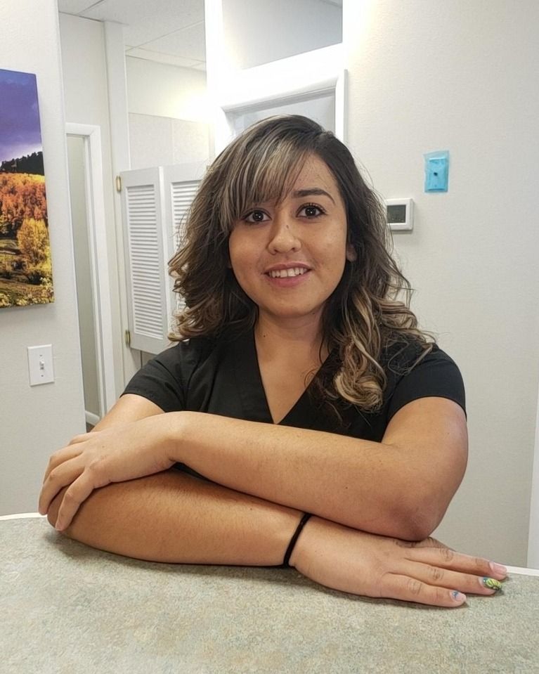 Woman with light brown hair smiles, arms crossed, leaning on counter in a clinic setting.