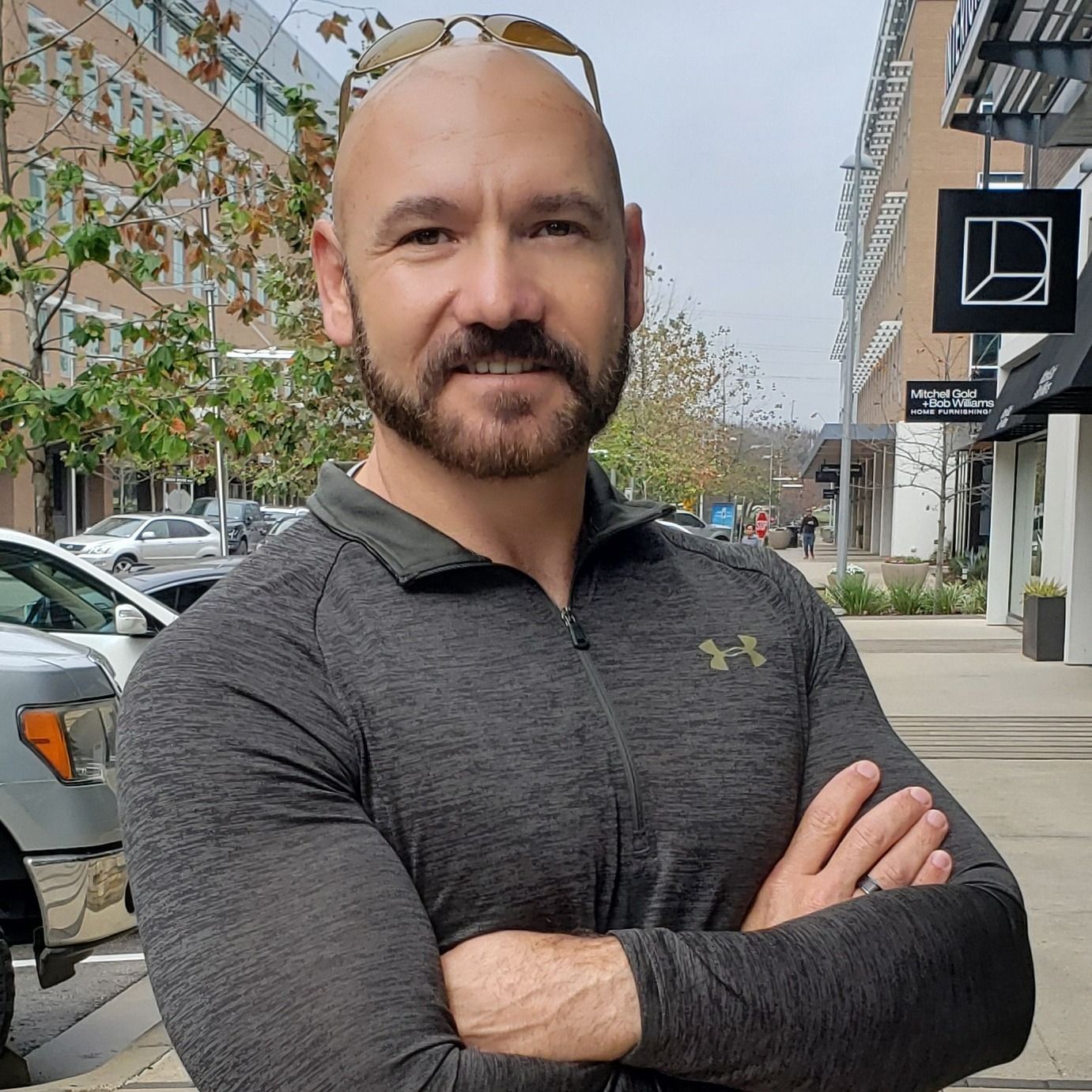 A bald person with a beard, wearing a gray athletic quarter-zip, stands with arms crossed on an urban sidewalk.