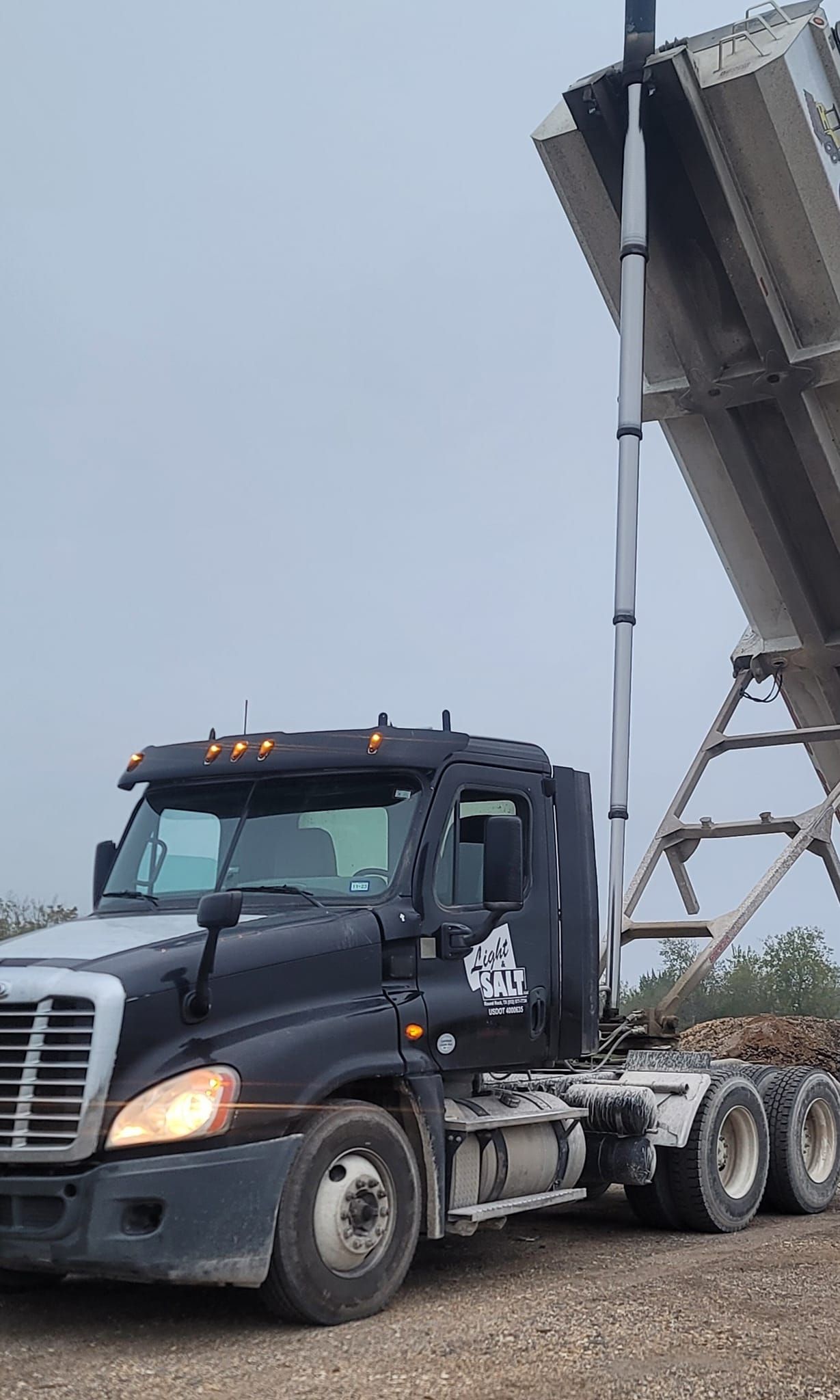 A dark Freightliner semi-truck with its dump trailer raised, parked on a gravel lot under a gray sky.