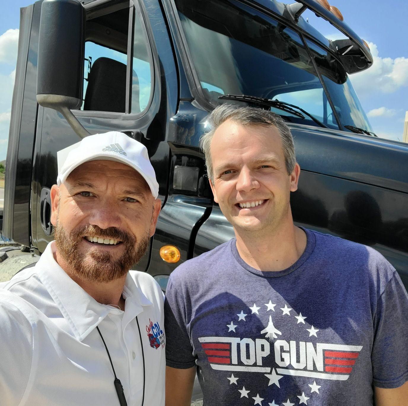 Two smiling men stand in front of a large black semi-truck on a sunny day. One wears a white cap, the other a Top Gun tee.