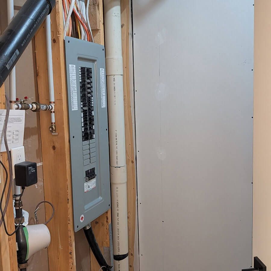 An electrical breaker panel mounted on wooden studs next to a white PVC pipe and a finished drywall wall.