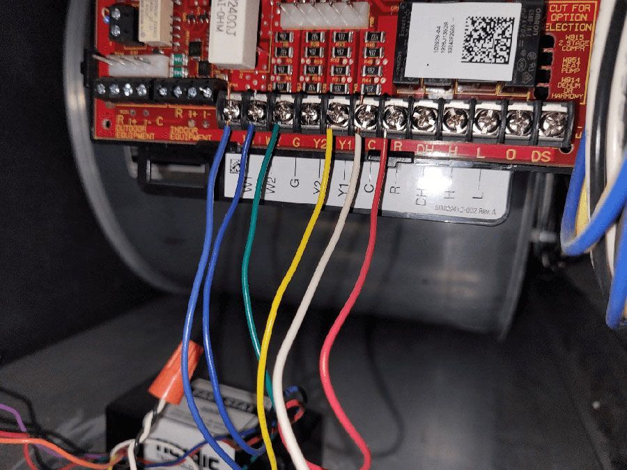 A close-up view of a red circuit control board for an HVAC system with blue, green, yellow, white, and red wires attached.
