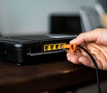 A person plugs an orange Ethernet cable into a yellow port on the back of a black network router.
