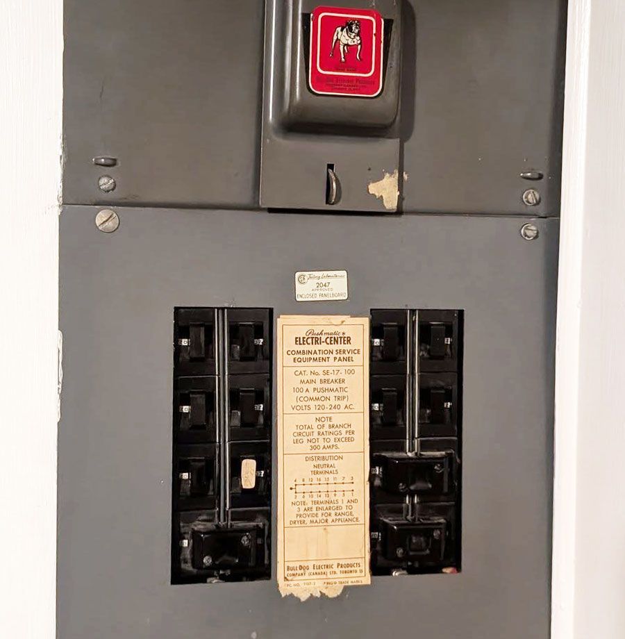 A gray electrical service panel mounted on a wall with a row of black circuit breakers and a central instructional label.