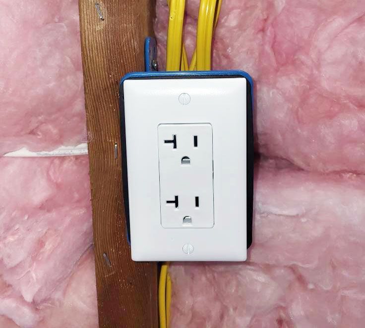 A 20-amp electrical outlet mounted on a wooden stud, surrounded by pink fiberglass insulation.