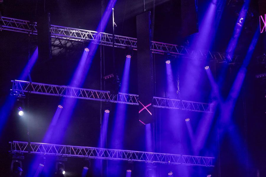 Stage lighting beams in vibrant blue cut through the darkness, illuminating metal truss structures hanging above.