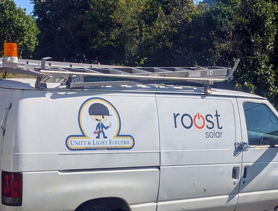 A white utility van with a ladder rack, featuring a Roost Solar logo and an Unity & Light Electric character graphic.