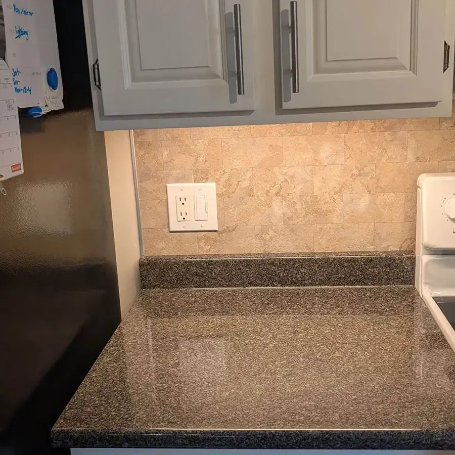 A kitchen counter with speckled grey stone, tan tile backsplash, white cabinets, and an electrical outlet.