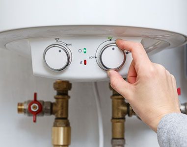 A hand adjusts the temperature dial on a white electric water heater control panel with brass plumbing fittings below.