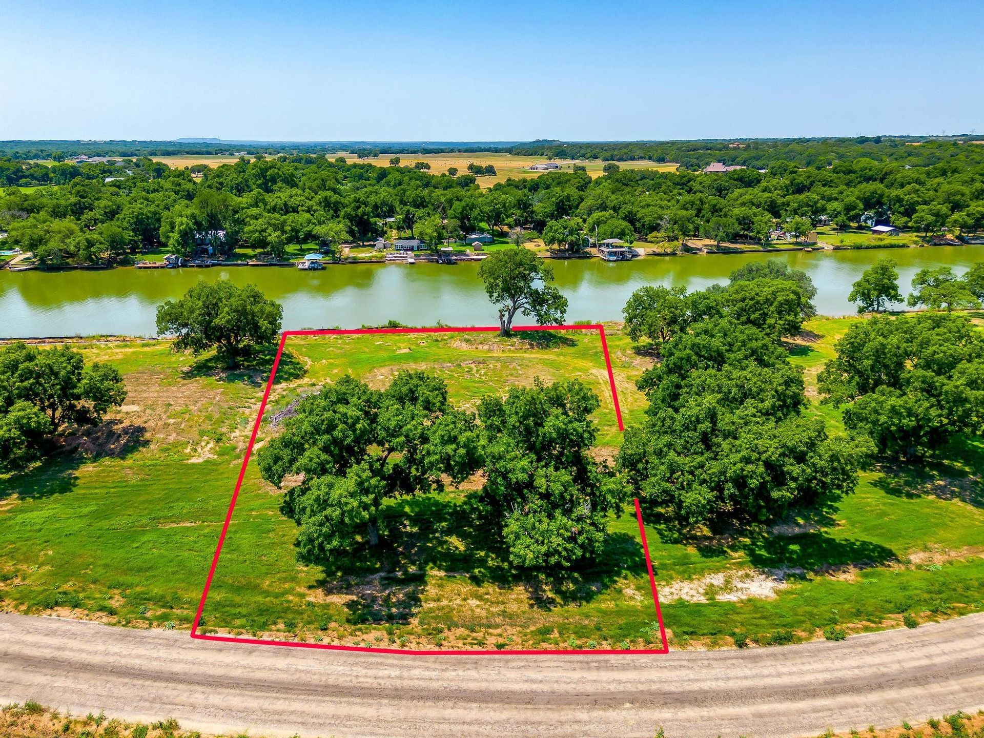 Green landscape with a river lined with trees, houses in the distance, and a dirt road.