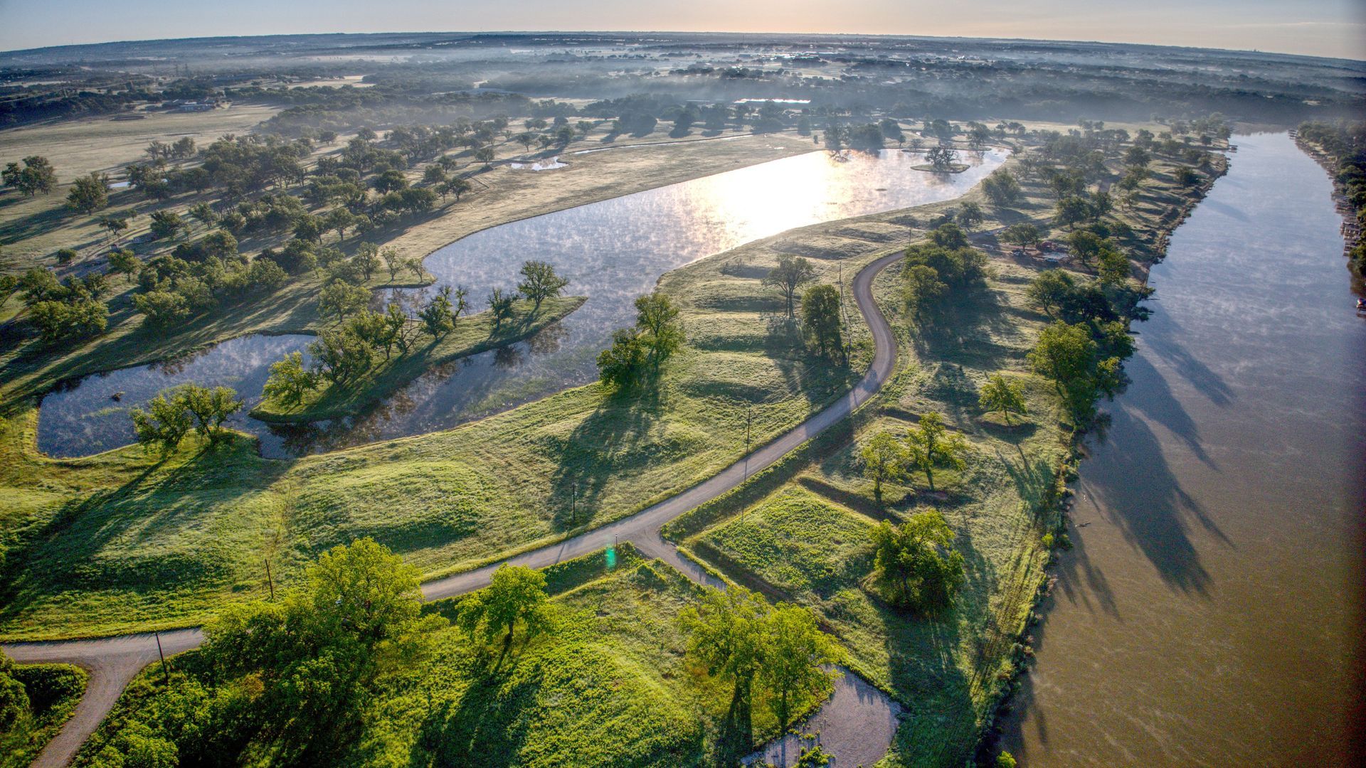 Aerial view of a green park with a river, ponds, trees, and a winding path.