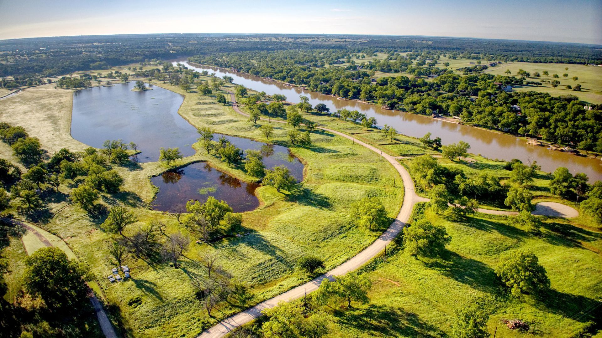 Aerial view of a green park with a river, ponds, trees, and a winding path.