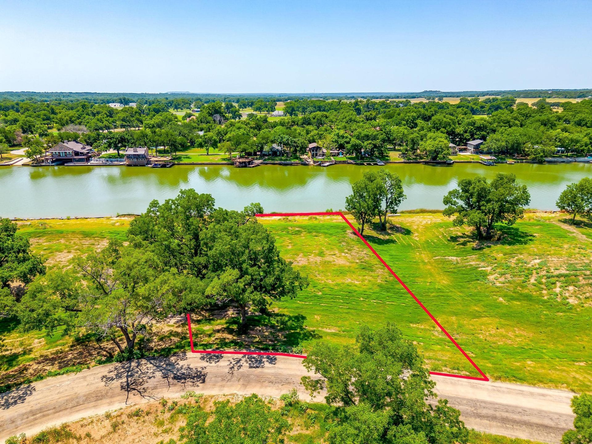 Aerial view of waterfront lot outlined in red, lush green grass and mature trees, houses along the water.