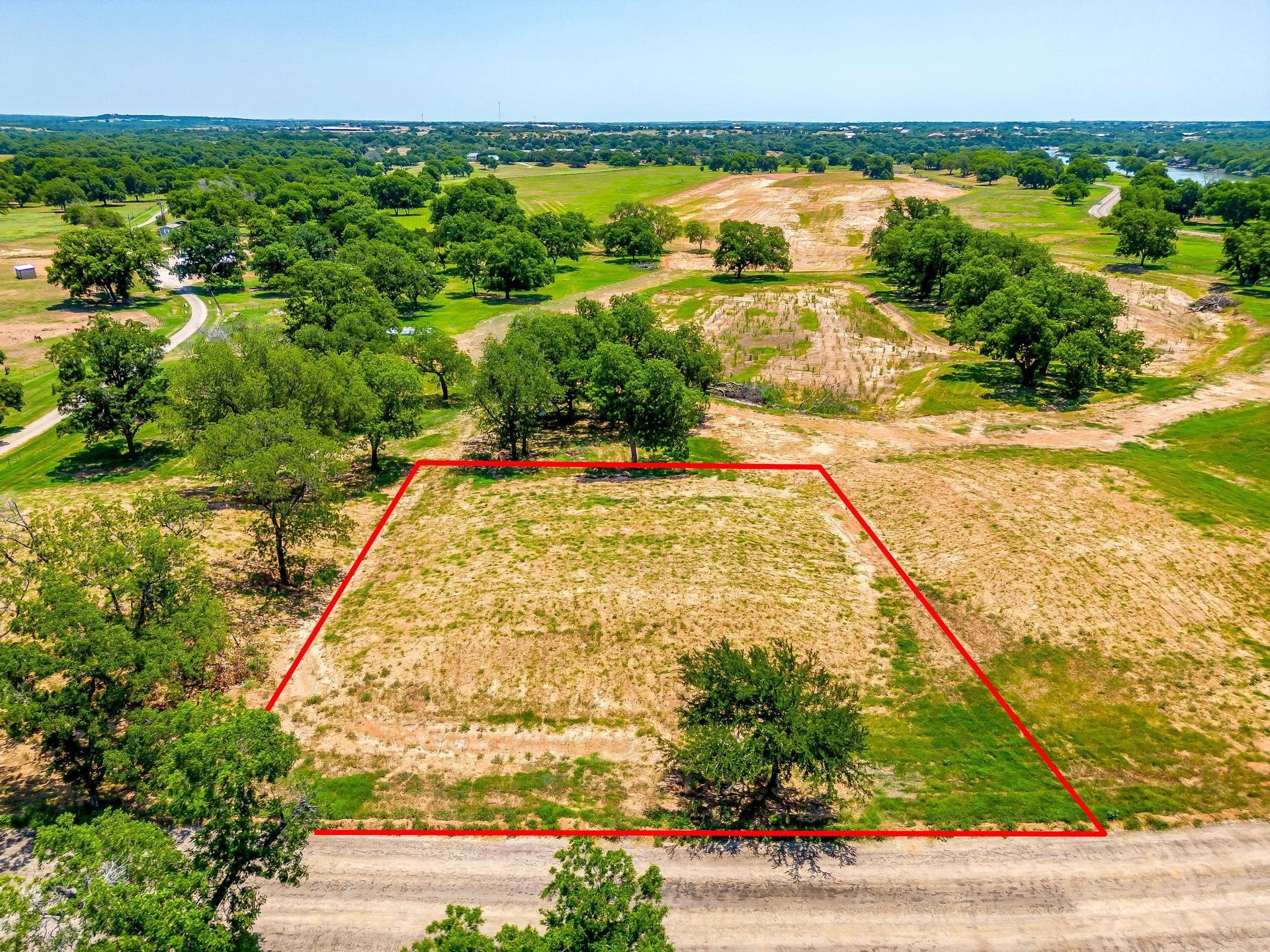 An aerial view of a cleared, grass-covered lot outlined in red, surrounded by trees and fields, near a road.