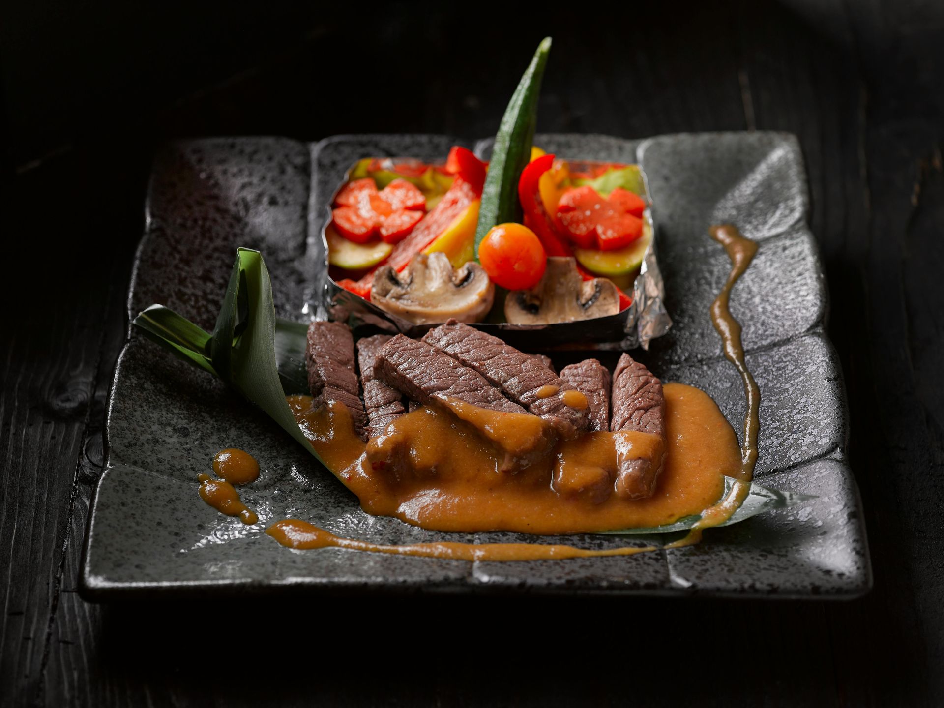 Sliced steak with sauce, alongside a colorful vegetable terrine on a dark square plate.